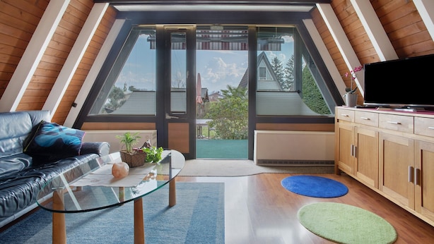 A modern loft bedroom with a sloped ceiling, a glass door leading to a balcony, a leather sofa, a glass coffee table, and a wooden cabinet.