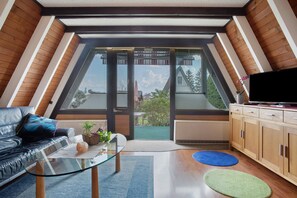 A modern loft bedroom with a sloped ceiling, a glass door leading to a balcony, a leather sofa, a glass coffee table, and a wooden cabinet. - Holiday Home 'Nurdach-Haus (Type 7)' with Private Terrace, Private Garden and Wi-Fi (Immenstaad am Bodensee)