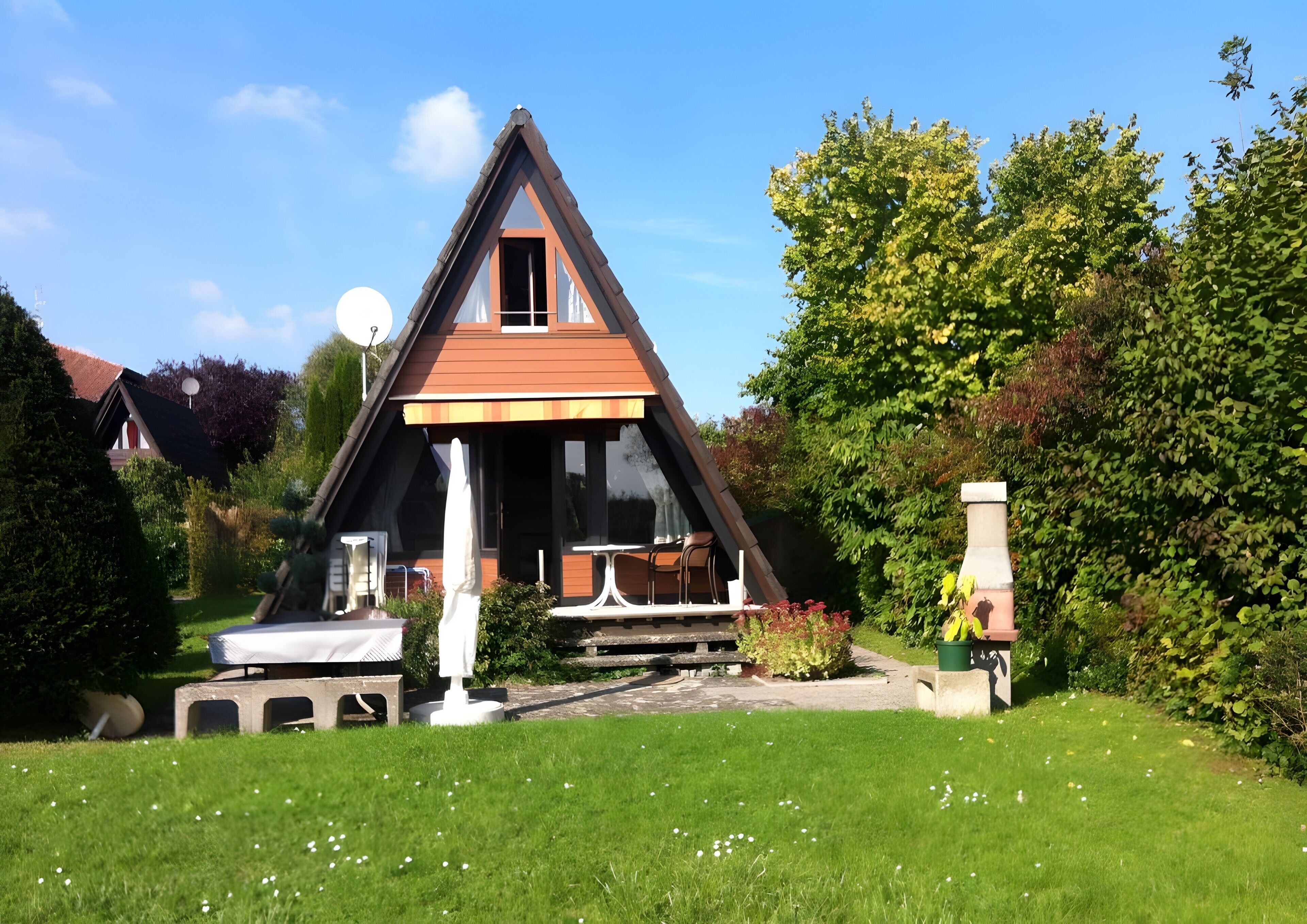 Une maison triangulaire avec une entrée bien visible, un petit patio aménagé avec du mobilier d’extérieur et un jardin agrémenté de fleurs blanches.