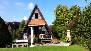 A triangular house with a visible entrance, a small patio area with outdoor furniture, and a garden with white flowers.