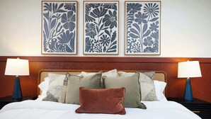 Une chambre d’hôtel comprenant un lit, des oreillers, deux lampes de chevet et trois œuvres d’art florales encadrées, disposées sur le mur.