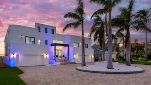 A modern two-story house with a paved driveway, palm trees, and a well-lit entrance. - Luxury Beachfront Retreat~Pool~Game Room~Sleeps 16 (Bradenton)