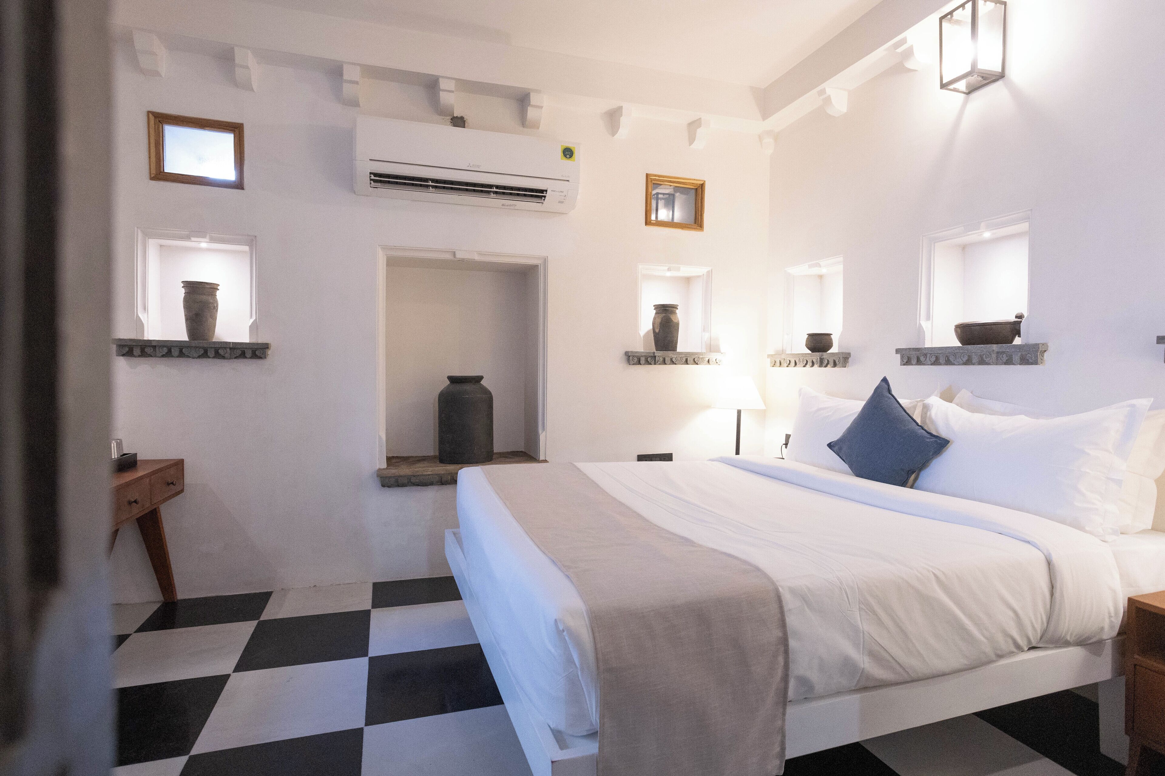 Habitación de hotel con piso a cuadros, una cama con ropa blanca, una mesita de noche y un aire acondicionado instalado en la pared, además de una pequeña ventana con un florero en el repisa.