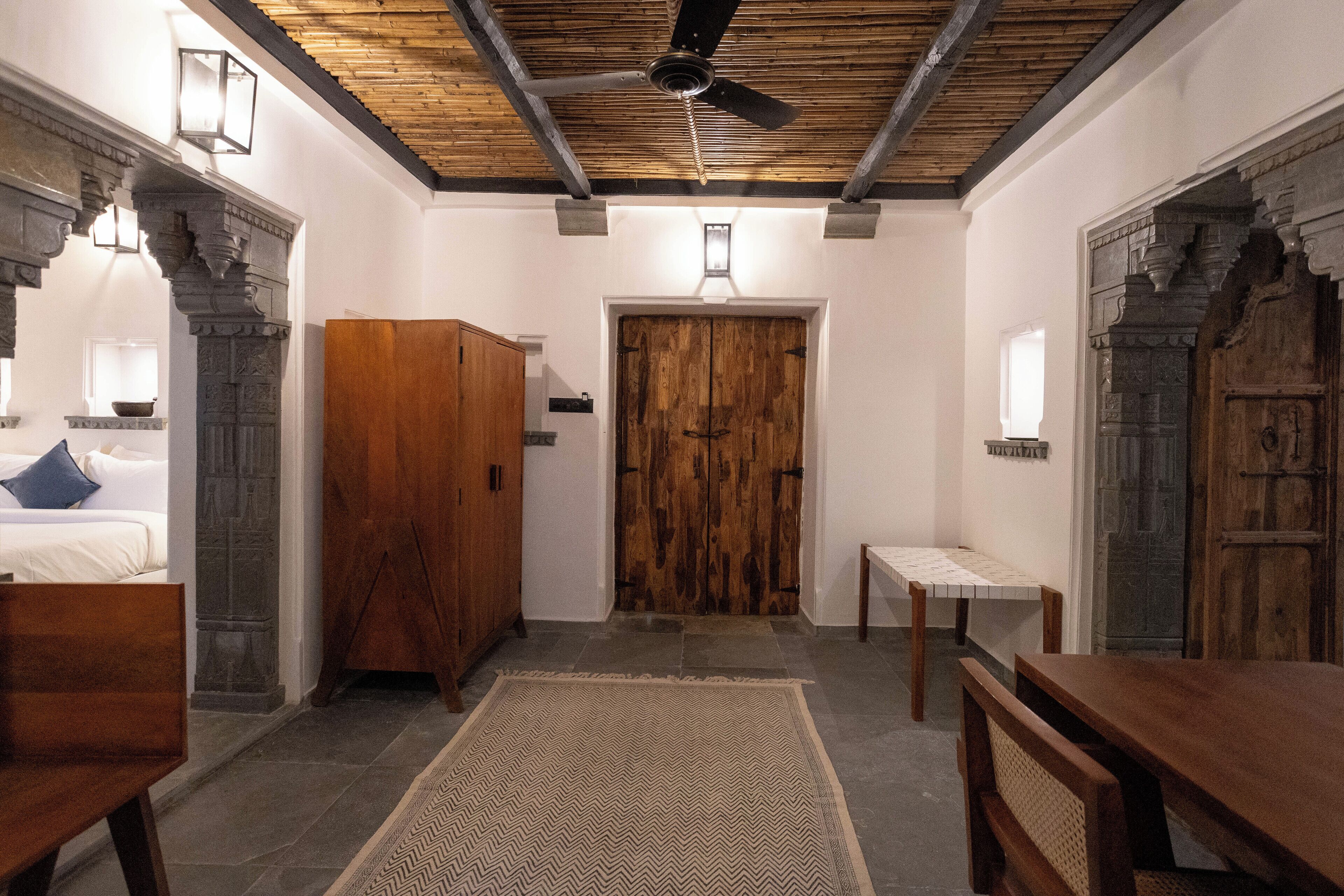 A room with a wooden ceiling, a large wooden door, a bed with white bedding, a wooden bench, and a small table.