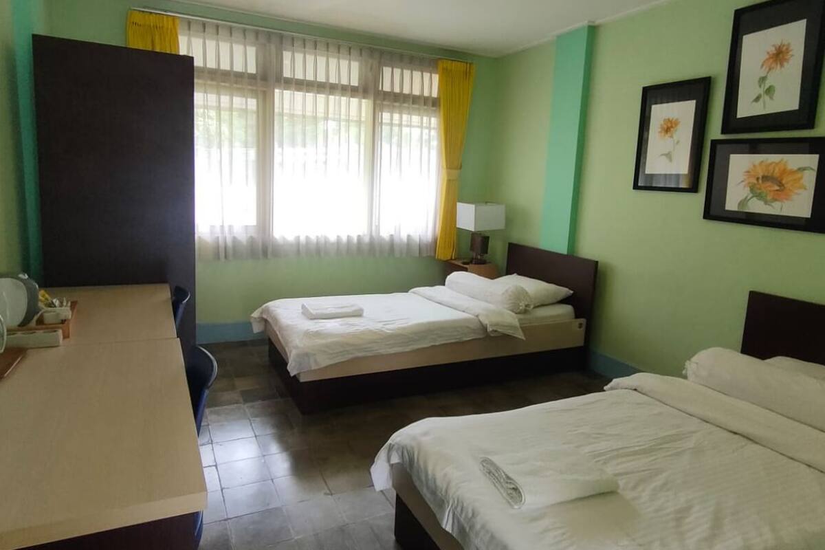Ein Hotelzimmer mit zwei Betten, einem Schreibtisch, einem Stuhl, einem Fenster mit Vorhängen und gerahmten Bildern an der Wand.