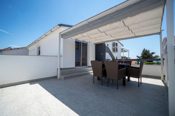 A covered outdoor patio with a table and chairs, a white building in the background, and a clear blue sky. - Villa Skundric (Vir)