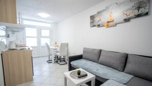 A modern living room with a grey sofa, a small white table with a remote control, and a kitchen area with a wooden cabinet and white chairs. - Villa Skundric (Vir)