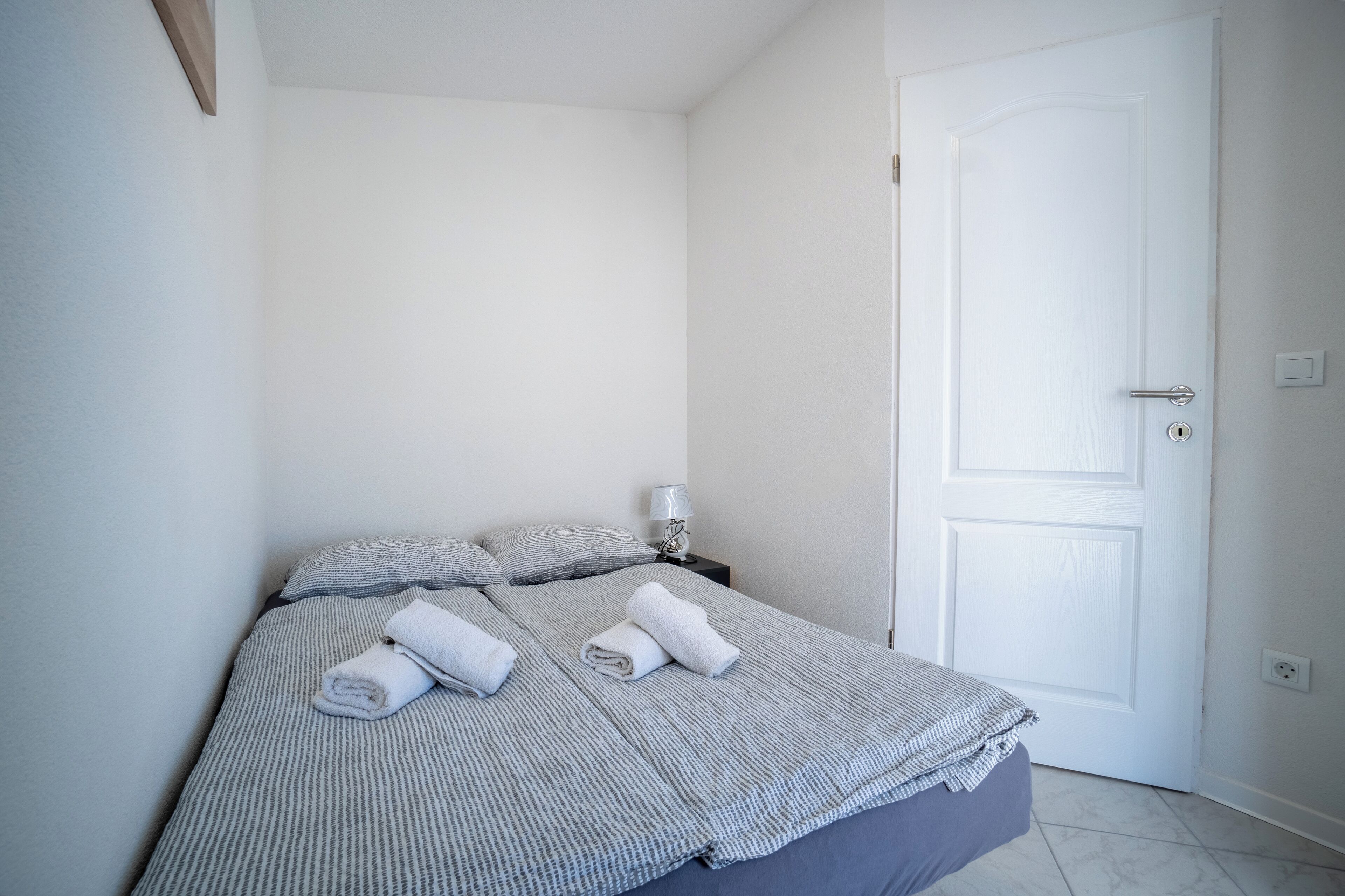 A neatly made bed with a striped bedspread and pillows, a bedside lamp, and a white door.
