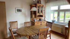 A dining area with a wooden table and chairs, a clock, and a window with a view of greenery. - Apartment 'Uhlenhoff Boyken' with Private Terrace, Shared Garden and Wi-Fi (Apen-Bokelermoor)