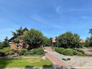 A brick house with a red roof, surrounded by green trees and a gravel pathway.