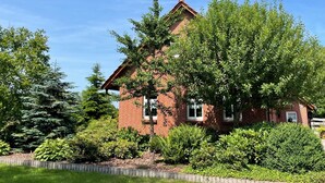 A brick house with a sloped roof, surrounded by various green shrubs and trees. - Apartment 'Uhlenhoff Boyken' with Private Terrace, Shared Garden and Wi-Fi (Apen-Bokelermoor)