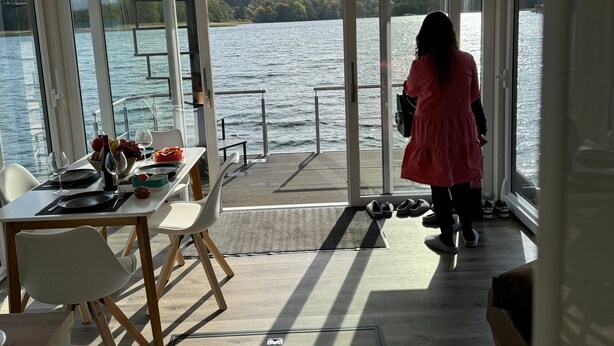 A dining area with a table set for two, a view of the lake, and a person standing by the window. - Houseboat 'Kitilou' with Lake View, Private Terrace, and Wi-Fi (Dobin)