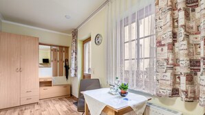 A room with a wooden wardrobe, a dining table with a white tablecloth, a chair, a potted plant, a radiator, and a window with patterned curtains.