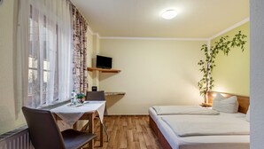 A small room with a bed, a desk with a chair, a TV on a shelf, and a plant.