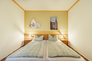 Una habitación con dos camas, un cabecero de madera, luces fijadas a la pared, un florero, platos y un cuadro de un edificio.