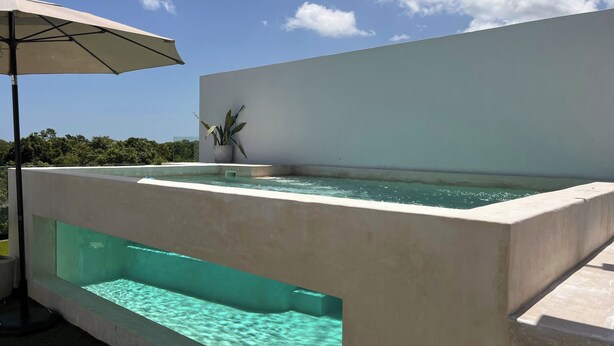Un área de piscina moderna al aire libre, con piscina azul cristalina, una planta en maceta y una pared blanca.