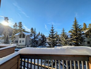A snow-covered deck with a wooden railing, a house with a sloped roof, and evergreen trees in a mountainous area. - Key Condo 2934a (Keystone)
