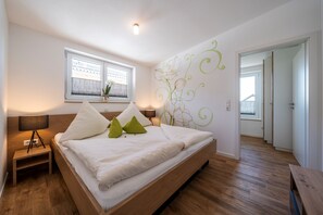 Une chambre à coucher avec un lit en bois, une table de chevet, une fenêtre donnant sur un beau paysage et un mur orné d’un motif floral.