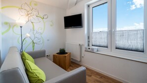 Un salon moderne avec un canapé, un téléviseur à écran plat fixé au mur, une fenêtre avec des stores et un radiateur.