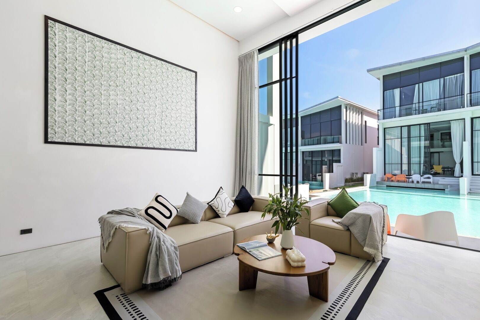 Una sala de estar moderna con un sofá color beige, una mesa de centro y un amplio ventanal con vistas a una piscina y edificios cercanos.