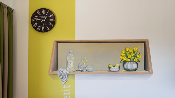 Une horloge murale, une étagère avec une bouteille, des verres, un bol de fruits et un vase de fleurs.