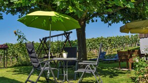 Une terrasse avec une table et des chaises sous un parasol vert, entourée d’arbres et d’un vignoble.