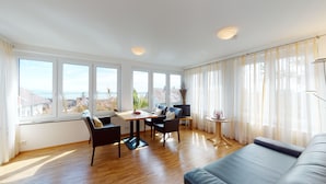 A modern living room with a wooden floor, a sofa, a dining table with chairs, a TV, and large windows with curtains.