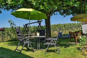A patio with a table and chairs under a green umbrella, surrounded by trees and a vineyard.
