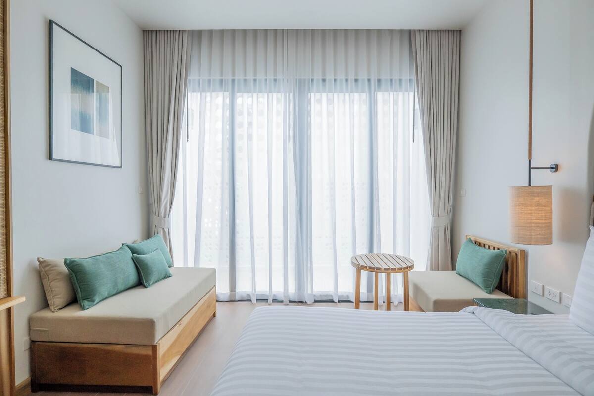 A hotel room with a large bed, a sofa, a small table, and a window with sheer curtains.