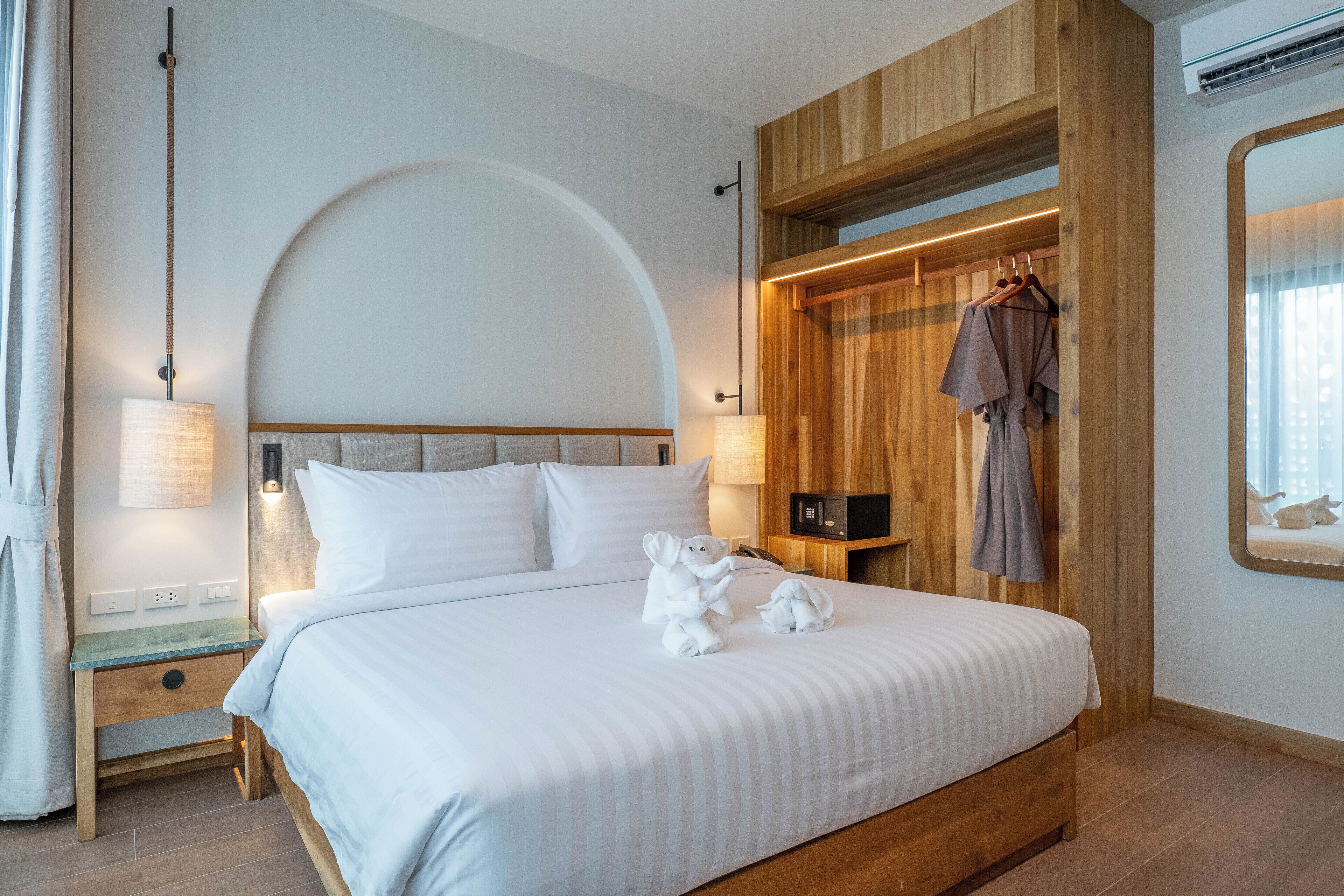 Quarto de hotel moderno com cama grande, cabeceira de madeira e roupeiro embutido com roupas penduradas.