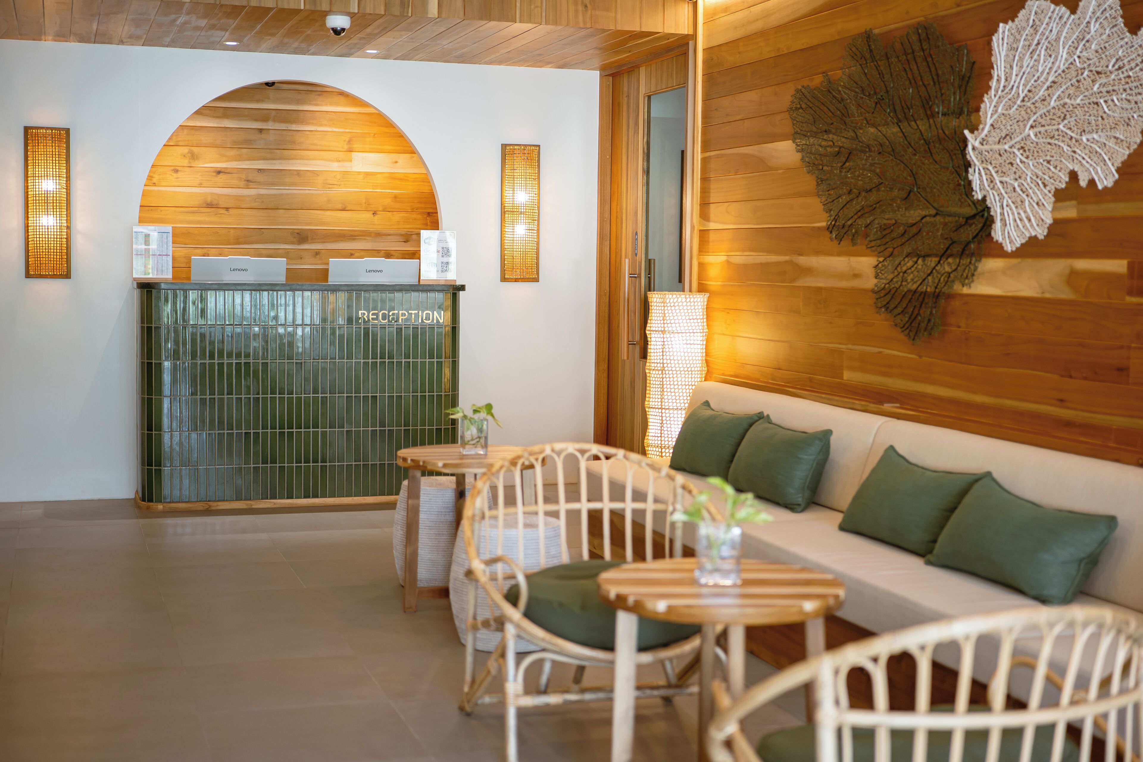 Un'area reception con una scrivania in legno, un bancone reception verde, sedie in vimini, un divano bianco con cuscini verdi e un pannello decorativo a forma di foglia.