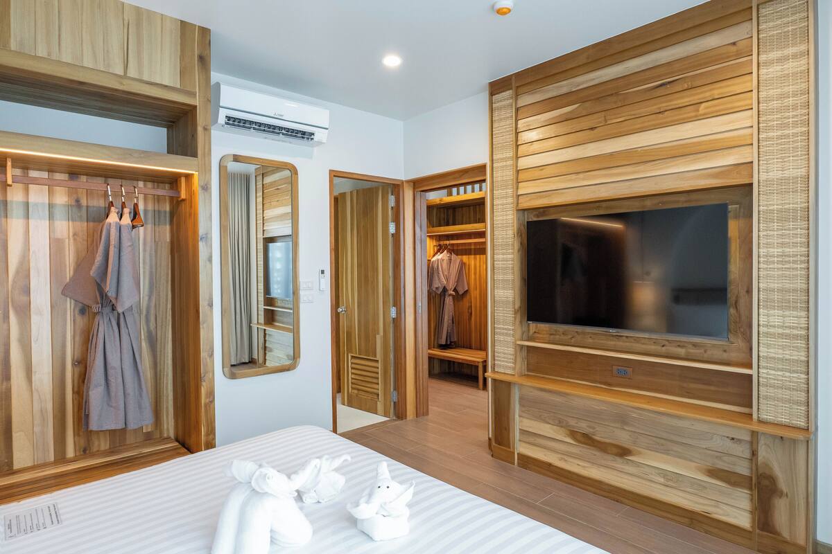 A hotel room with wooden paneling, a large flat-screen TV, a bed with white linens, and a walk-in closet with hanging clothes.