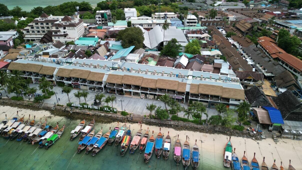An aerial view of a coastal area with a row of boats on the beach and a building with a thatched roof in the background.