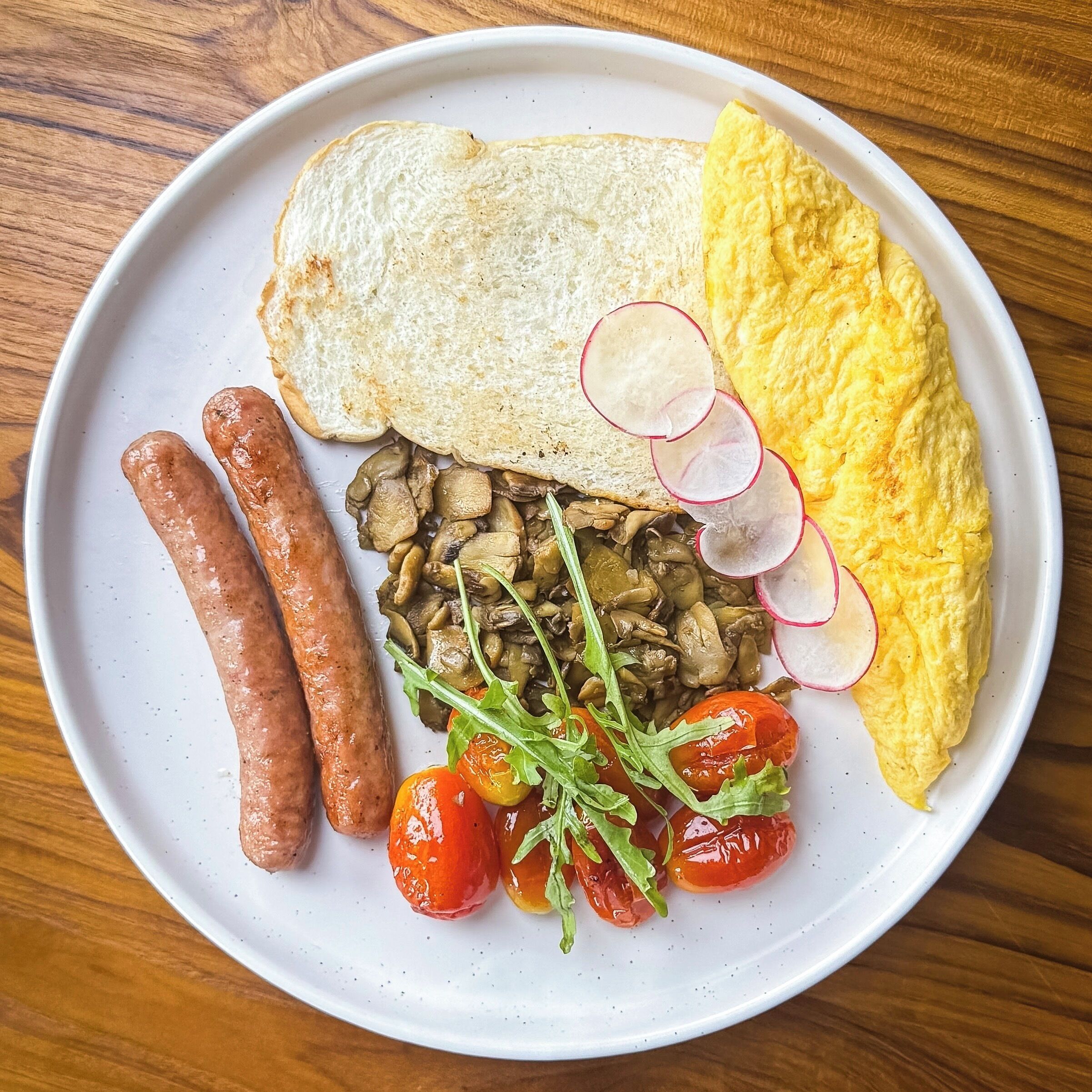 Un piatto con pane tostato, uova strapazzate, salsicce, funghi, ravanelli, pomodorini e rucola.