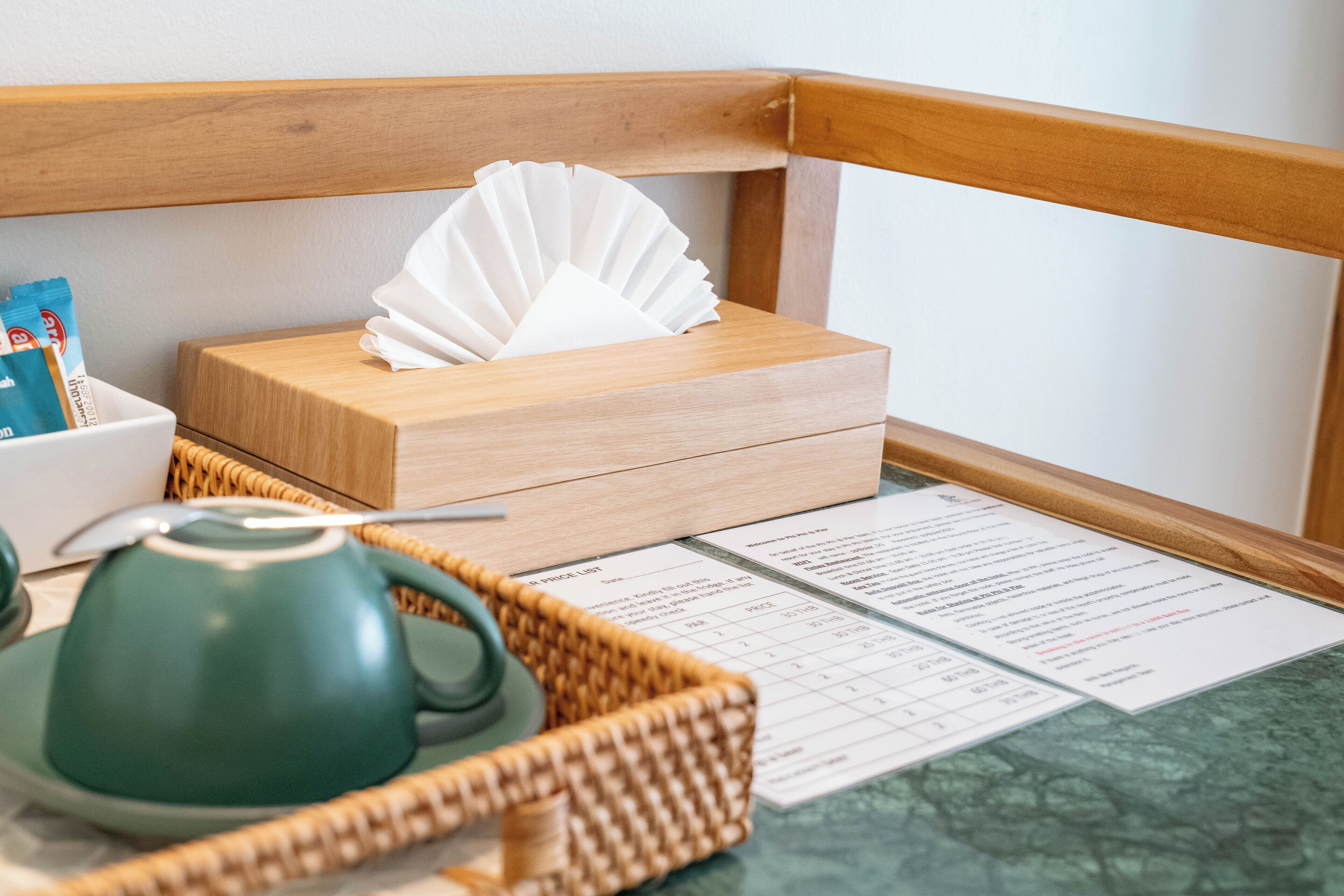 A wooden box with a tissue, a green teapot, a spoon, a basket, and a menu on a table.