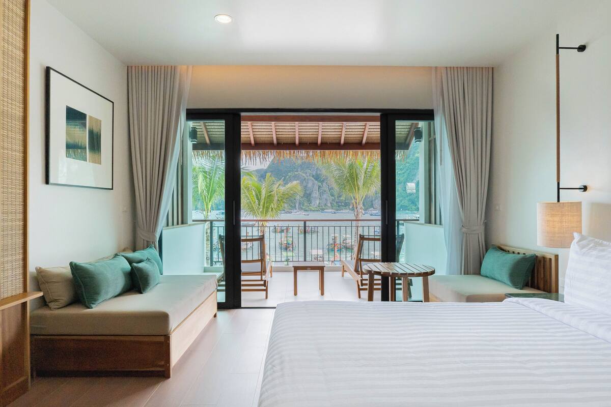 A hotel room with a large bed, a sofa, a small table, and a balcony with a view of palm trees and boats.