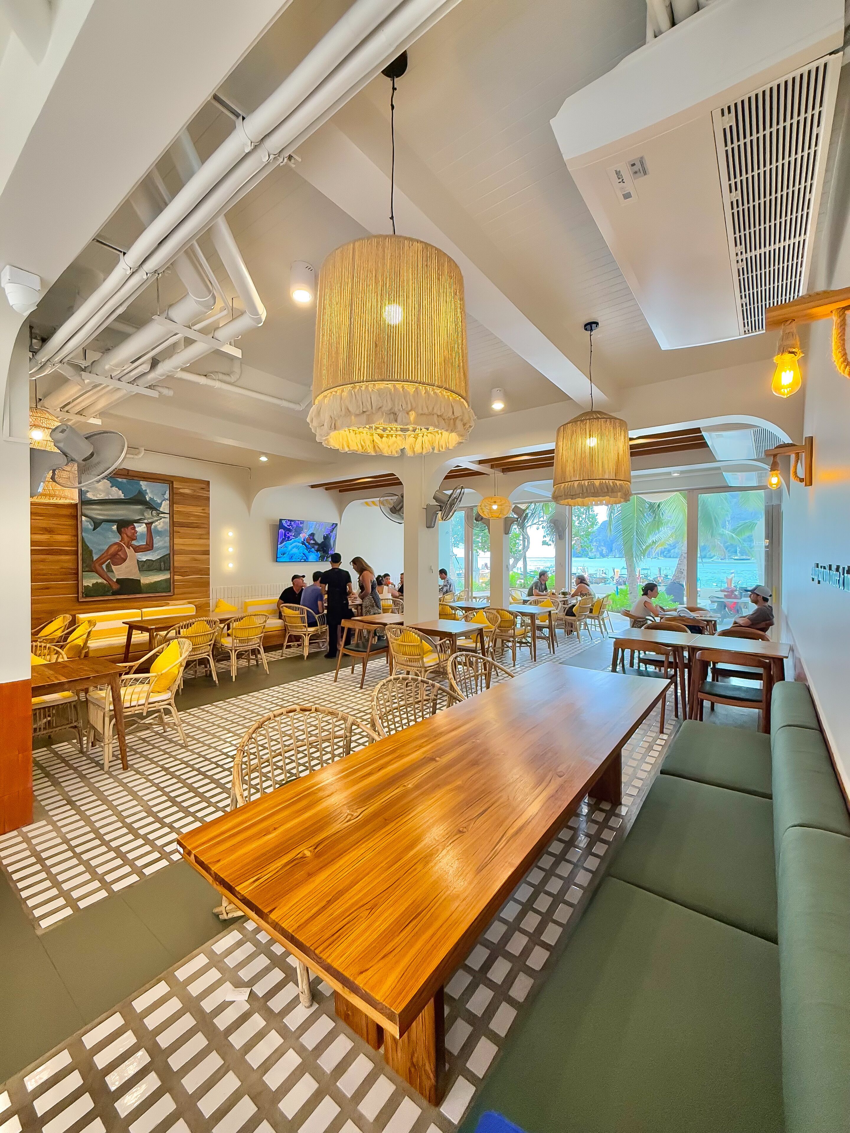 Un restaurante moderno con mesas de madera y asientos acolchados verdes, grandes lámparas colgantes y vistas al exterior.