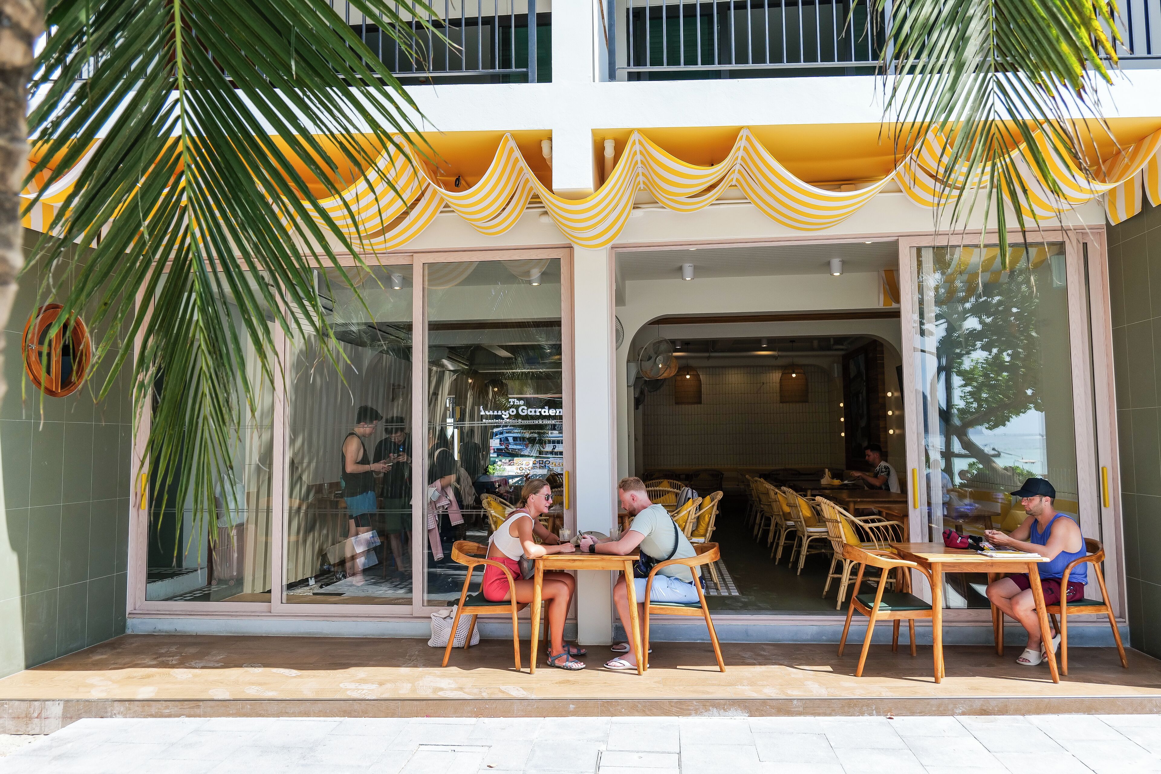 An outdoor seating area with wooden tables and chairs, a palm tree, and a building entrance with large windows and yellow-striped awnings.