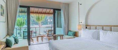 A hotel room with a large bed, a sofa, a small table, and a balcony with a view of greenery.