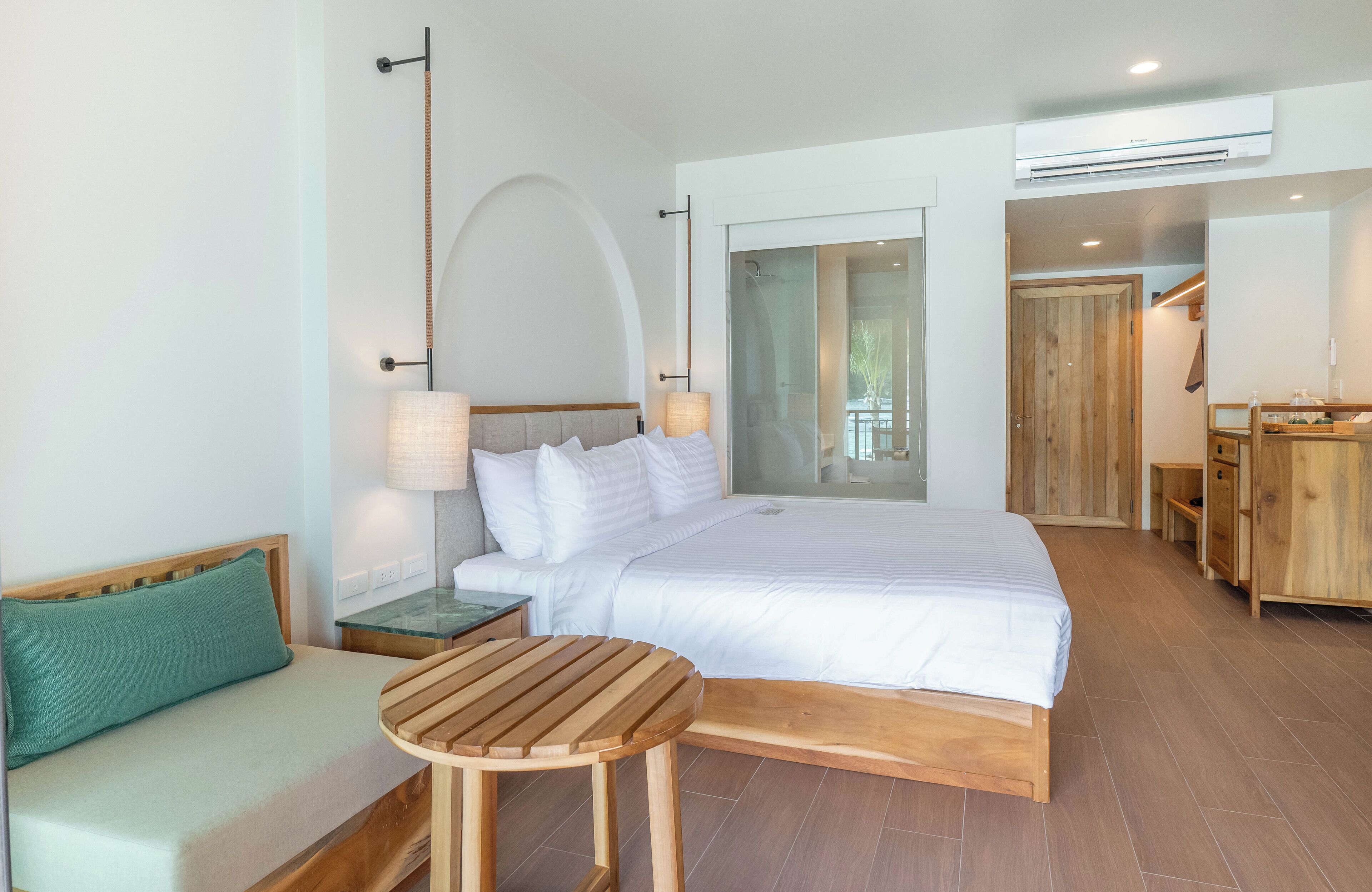 Un dormitorio moderno con una cama grande, un banco de madera, una mesita pequeña y una zona de ducha de vidrio.