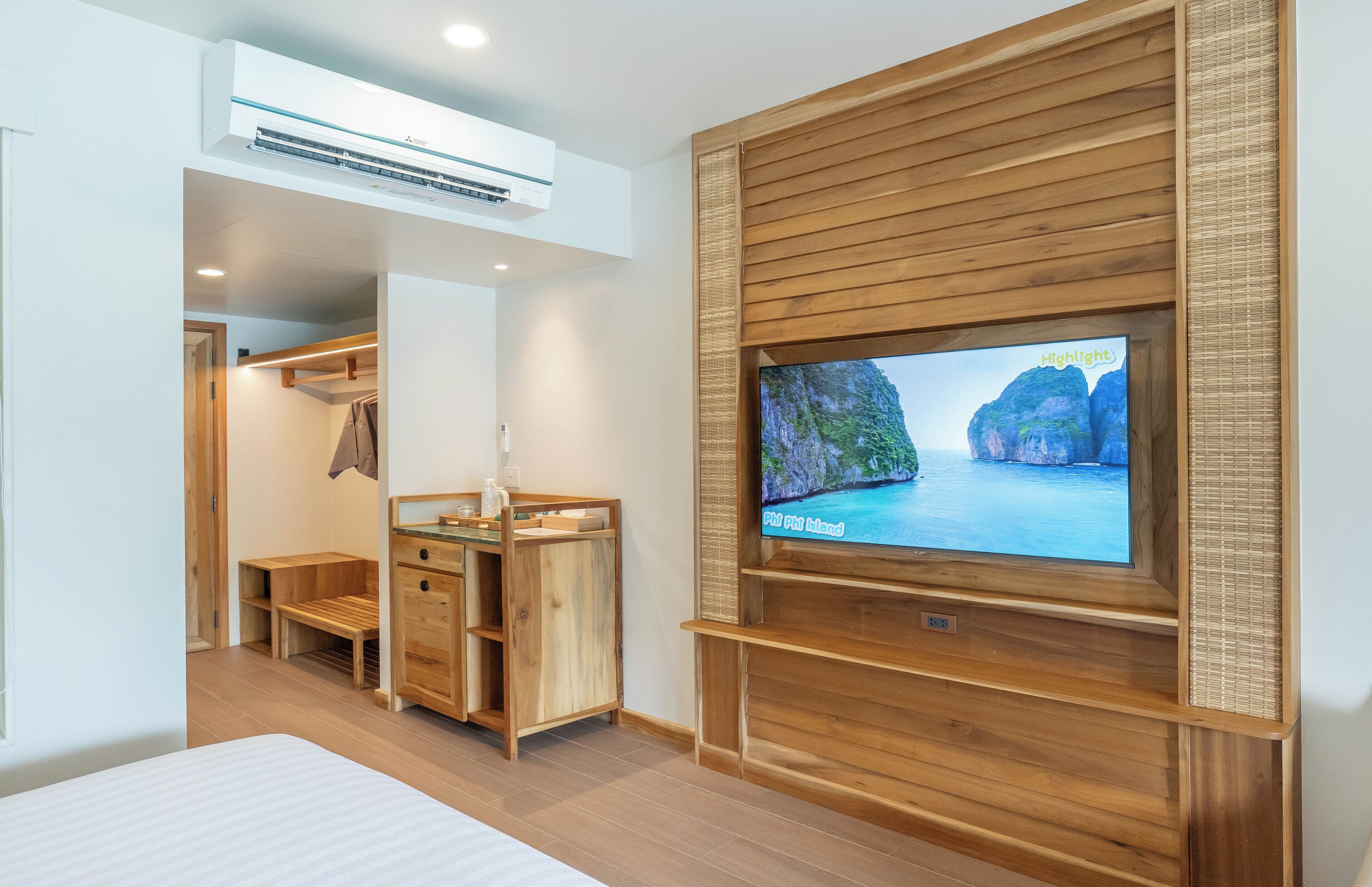 A modern hotel room with a wooden TV stand, a flat-screen TV displaying a scenic view, a wooden wardrobe, and a small wooden bench.