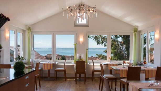 A spacious dining area with multiple tables and chairs, large windows with ocean views, and a chandelier. - Room '102' with Shared Garden, Balcony and Wi-Fi (Hagnau)