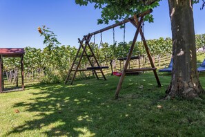 Un terrain de jeux avec des balançoires et un arbre, entouré d’une pelouse et avec un vignoble en arrière-plan.