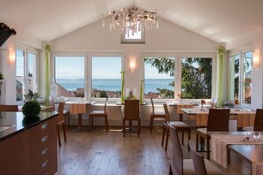 Un comedor espacioso con varias mesas y sillas, amplias ventanas con vistas al océano y un candelabro.