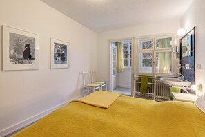 Une chambre à coucher comprenant un lit, deux tableaux encadrés, une fenêtre, un radiateur, ainsi qu’une salle de bain avec un lavabo et un miroir.