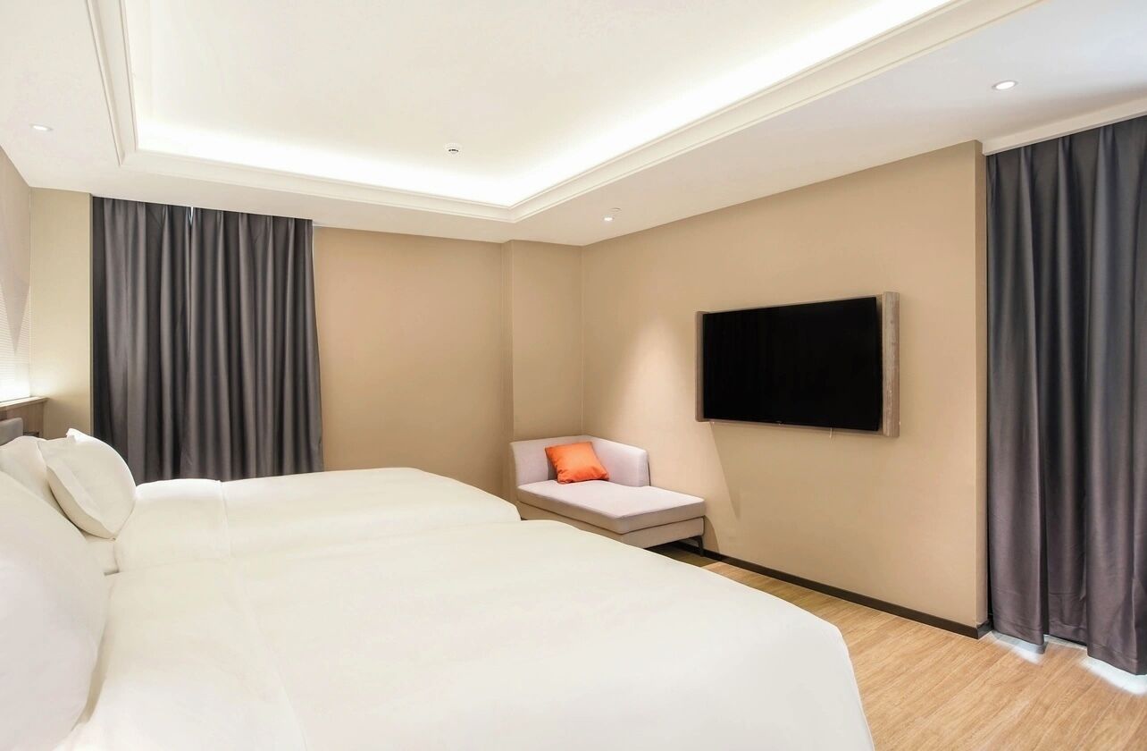 A hotel room with a large bed, a small sofa with an orange pillow, a flat-screen TV mounted on the wall, and dark curtains on the windows.
