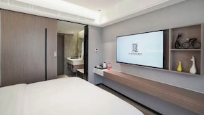 Una habitación de hotel moderna con una cama, un televisor montado en la pared, un baño visible a través de una puerta abierta y una repisa con objetos decorativos.