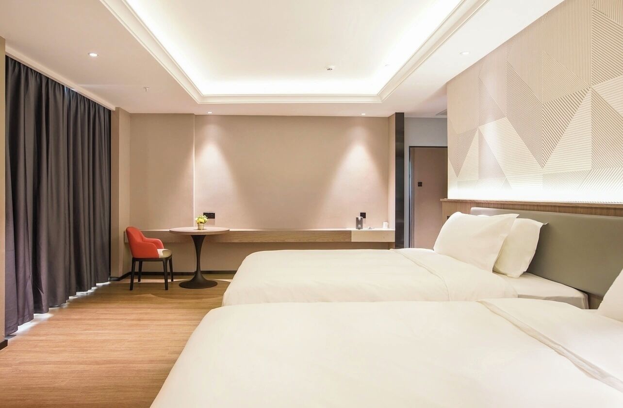 A hotel room with two beds, a small table with a chair, a large window with curtains, and a geometric wall design.