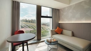 Una habitación de hotel moderna con un ventanal grande, un sofá, una mesita y una silla roja.
