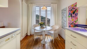 Una habitación con una mesa redonda, sillas blancas, una lámpara colgante, un radiador y una ventana con cortinas.